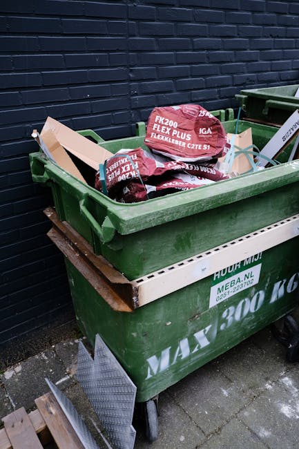 A large green wheeled dumpster filled with red and black bags of building materials, positioned against a dark blue brick wall outdoors on a paved surface. The bags are labeled with technical specific