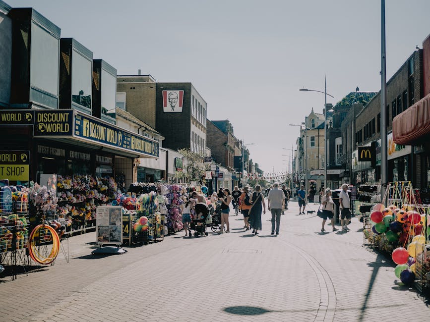 A busy pedestrian street in an urban shopping area during daytime, with numerous people walking and browsing the storefronts. On the left side, outdoor stalls display an assortment of colorful merchan