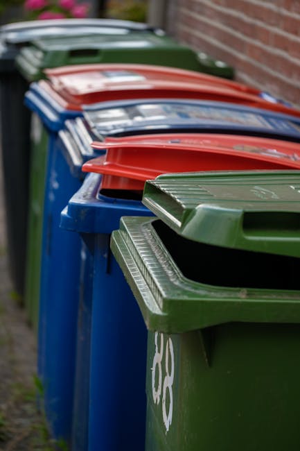 A person's hand and part of their forearm are visible in the image, holding a green and blue plastic waste bag by its twisted top. The bag is semi-transparent, showing some of the contents inside, and