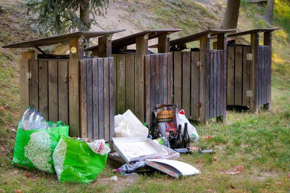 Four wooden outdoor storage units with sloped roofs are positioned side by side on a grassy area near a wooded background, each with visible hinges and locks. In front of the units, there is an assort
